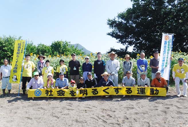 ひまわりを背景に社会を明るくする運動の横断幕をもつ運動の参加者数十名による集合写真